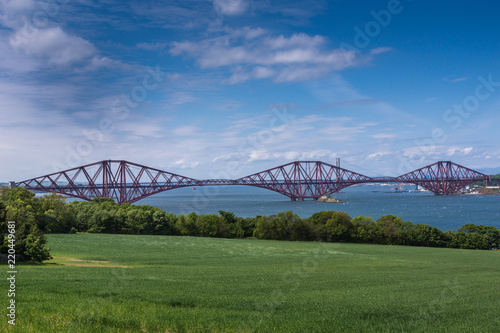 Queensferry, Scotland, UK - June 14, 2012: Red metal iconic Forth Bridge for trains over Firth of Forth under blue sky. Green field up front. © Klodien