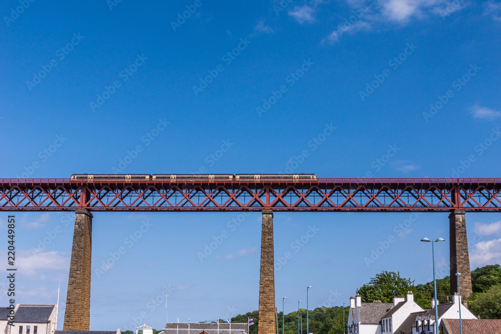 Queensferry, Scotland, UK - June 14, 2012: Train rides over Red metal iconic Forth Bridge over Firth of Forth against blue sky. Tall supporting pillars and rooftops of the town. Queensferry, Scotland, UK - June 14, 2012: Train rides over Red metal iconic Forth Bridge over Firth of Forth against blue sky. Tall supporting pillars and rooftops of the town.