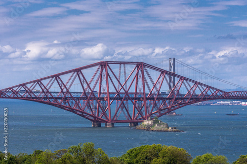 Queensferry, Scotland, UK - June 14, 2012: Closeup of One of three segments of Red metal iconic Forth Bridge over Firth of Forth between blue sky and water. Buildings on other side of water.  © Klodien