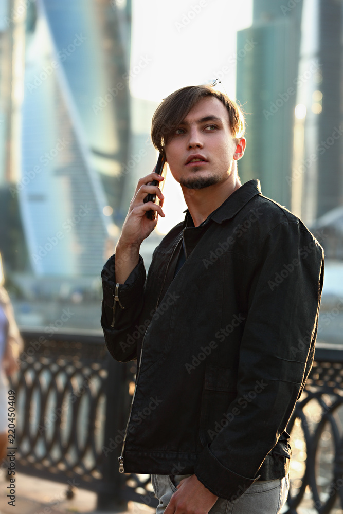 Fototapeta premium Young thinking casual man walking on the street and talking on mobile phone on city center background
