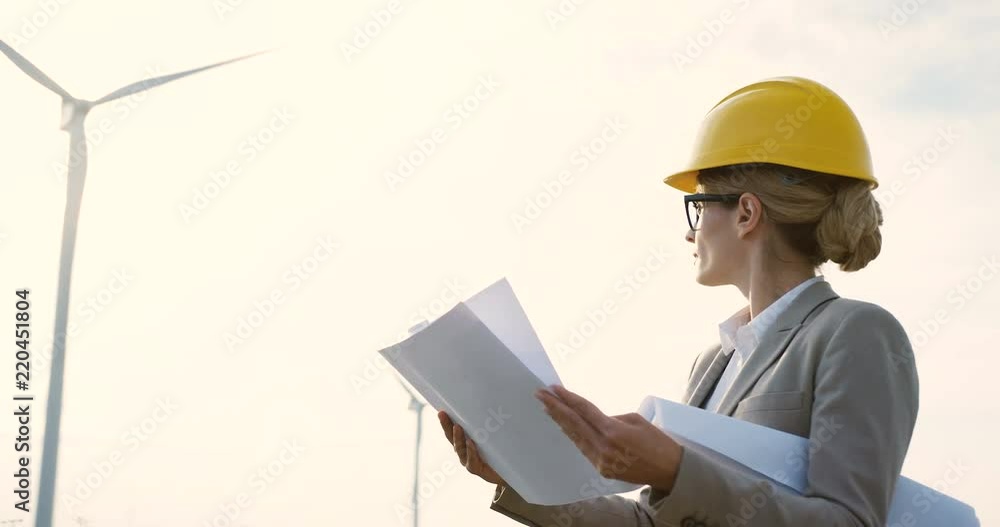 Portrait shot the woman in a helmet, environmental and safe power engineer, standing with some documents while watching the work of the big windmill turbine. Portrait shot the woman in a helmet, environmental and safe power engineer, standing with some documents while watching the work of the big windmill turbine.
