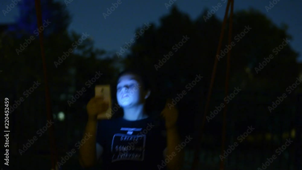 Girl using smart phone while riding on swing in nigth park. Selective focus. Urban people lifestyle background. Girl using smart phone while riding on swing in nigth park. Selective focus. Urban people lifestyle background.