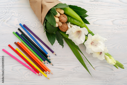 Fototapeta Naklejka Na Ścianę i Meble -  Small modest bouquet consisting of gladiolus, lemon, nuts and colour pencils on a white wooden table as a gift to the school child at the beginning of the school year
