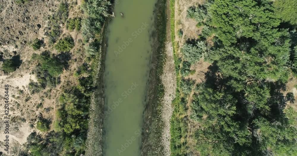 Aerial slow forward flight top to bottom of the Rio Grande in New Mexico. The river appears green and has rocky banks as well as green brush and trees. Aerial slow forward flight top to bottom of the Rio Grande in New Mexico. The river appears green and has rocky banks as well as green brush and trees.