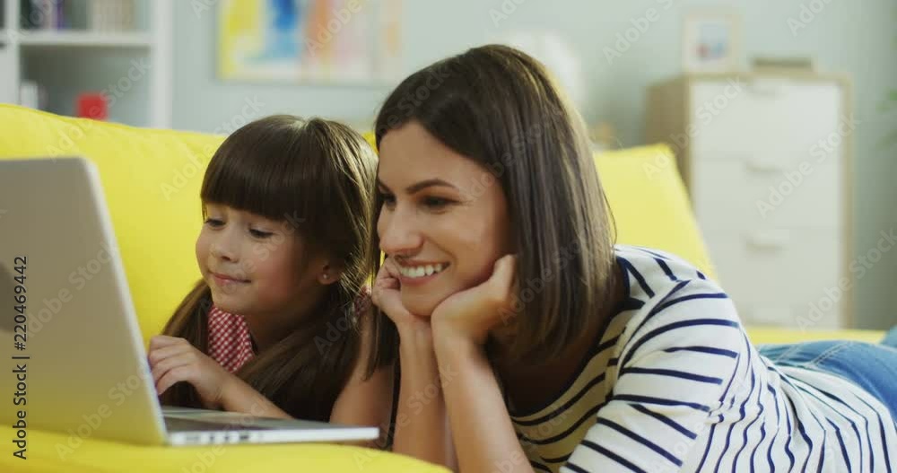 Close up of the good looking smiled mother and daughter watching something on the laptop and lying on the yellow sofa in the living room. Inside. Close up of the good looking smiled mother and daughter watching something on the laptop and lying on the yellow sofa in the living room. Inside.