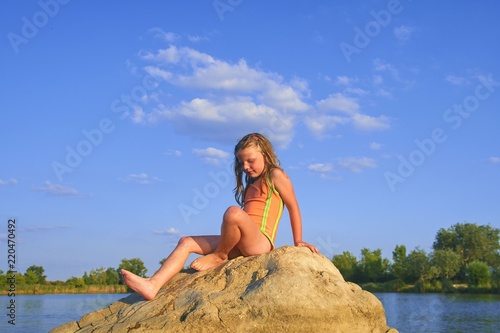 Beautiful girl sitting on a big rock. Little girl is wearing swimsuit. Summer and happy childhood concept. Copy space in bright blue sky © romannerud
