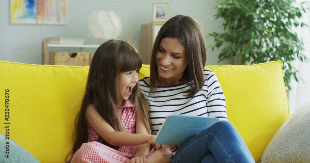 Portrait shot of the beautiful cheerful mother and pretty small daughter sitting together on the yellow couch and watching something on the tablet device in the living room. Portrait shot of the beautiful cheerful mother and pretty small daughter sitting together on the yellow couch and watching something on the tablet device in the living room.