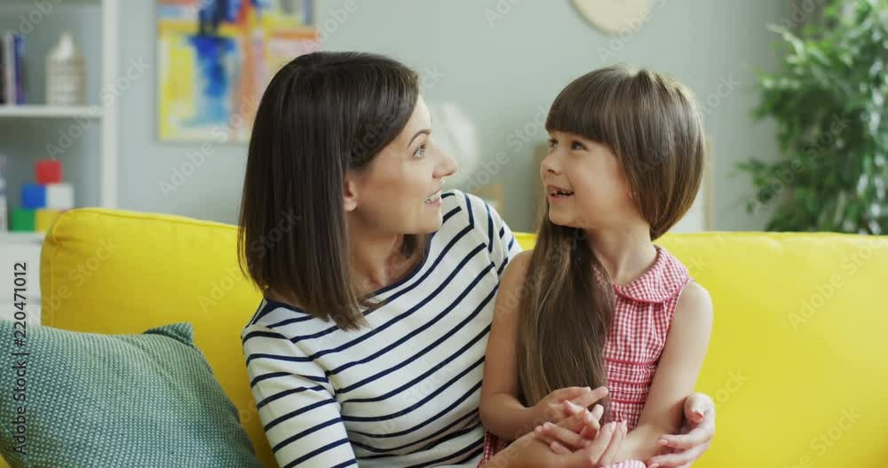 Portrait of the smiled beautiful young mother sitting on the yellow sofa in the living room and talking cheerfully with her little cute daughter who sitting on mom's knees at home. Portrait of the smiled beautiful young mother sitting on the yellow sofa in the living room and talking cheerfully with her little cute daughter who sitting on mom's knees at home.
