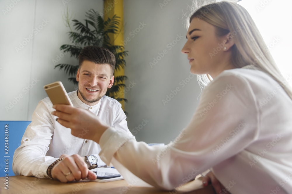 Girl is sitting at table with man and holding phone in hand. She is showing something to him. He is looking at phone and smiling. They are working together.