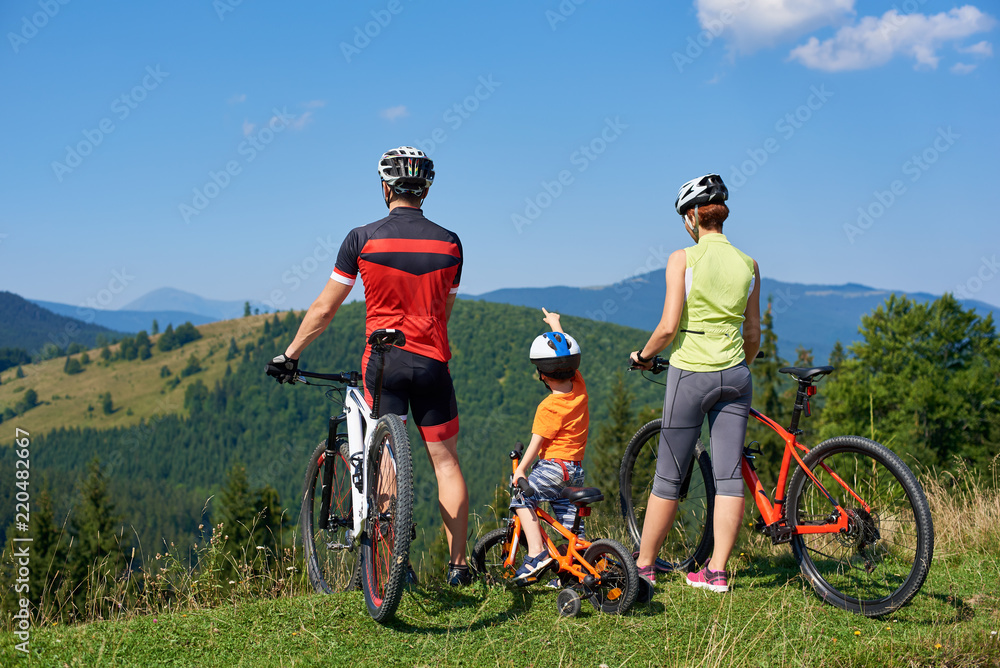 Naklejka premium Back view of tourist family bikers, mom, dad and child standing with bikes on top of grassy hill, enjoying beautiful mountains view. Boy pointing into a distance. Active lifestyle, traveling concept