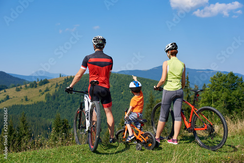 Back view of tourist family bikers, mom, dad and child standing with bikes on top of grassy hill, enjoying beautiful mountains view. Boy pointing into a distance. Active lifestyle, traveling concept