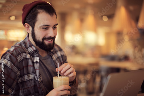 Handsome trendy hipster with glass of coffee drink using laptop in cafe