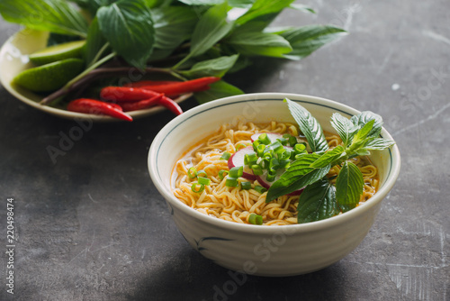Instant noodles in bowl with fresh herbs, garnish of cilantro and Asian basil, lemon, lime on dark stone background