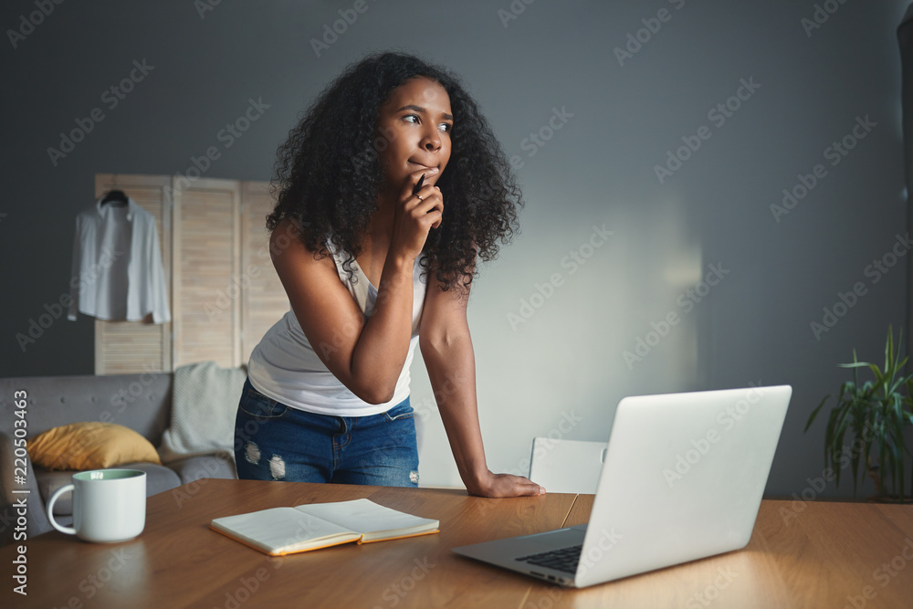 Picture of thoughtful dark skinned student girl in tank top and jeans touching chin with pensive facial expression, holding pen while writing essay, doing homework, standing in front of open laptop Picture of thoughtful dark skinned student girl in tank top and jeans touching chin with pensive facial expression, holding pen while writing essay, doing homework, standing in front of open laptop