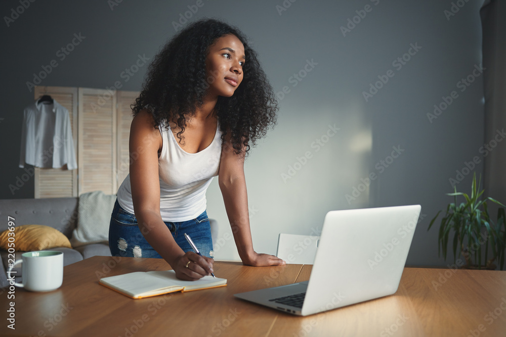 Stylish young African American woman journalist with curly hair standing at desk with open laptop and writing down in copybook, making research for new article. People, occupation and technology Stylish young African American woman journalist with curly hair standing at desk with open laptop and writing down in copybook, making research for new article. People, occupation and technology