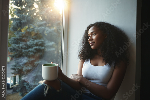 Portrait of beautiful twenty year old African American female wearing tank top and ripped jeans enjoying urban view while sitting on windowsill with mug of hot drink, feeling cozy and relaxed