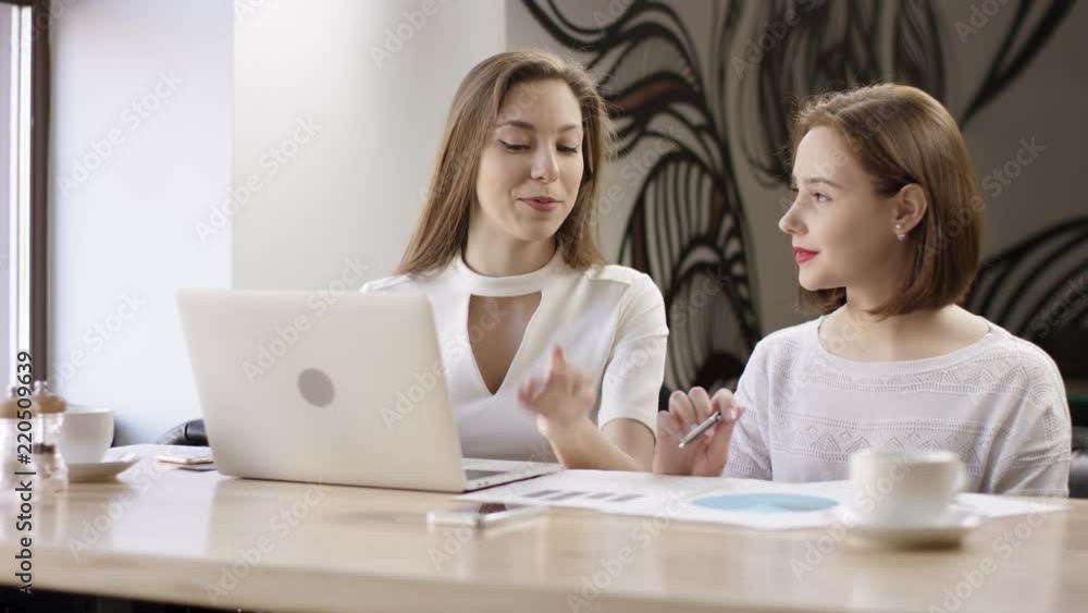 Two young business women are working with a laptop in a cafe. They are discussing and preparing the contract. It is a business meeting. Girls are sitting in the cafe at the table during cofee break Two young business women are working with a laptop in a cafe. They are discussing and preparing the contract. It is a business meeting. Girls are sitting in the cafe at the table during cofee break