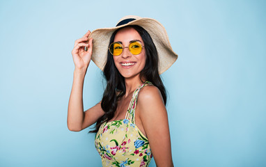 Portrait of cute summer brunette woman in hat, sunglasses and colorful dress, stylish girl have a fun and posing on blue background