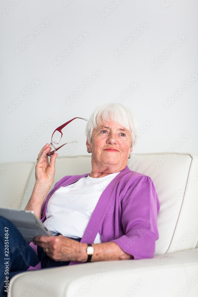 Portrait of a senior woman at home using a tablet computer - Looking happy, looking at the camera, smiling while sitting on the sofa in her living room Portrait of a senior woman at home using a tablet computer - Looking happy, looking at the camera, smiling while sitting on the sofa in her living room