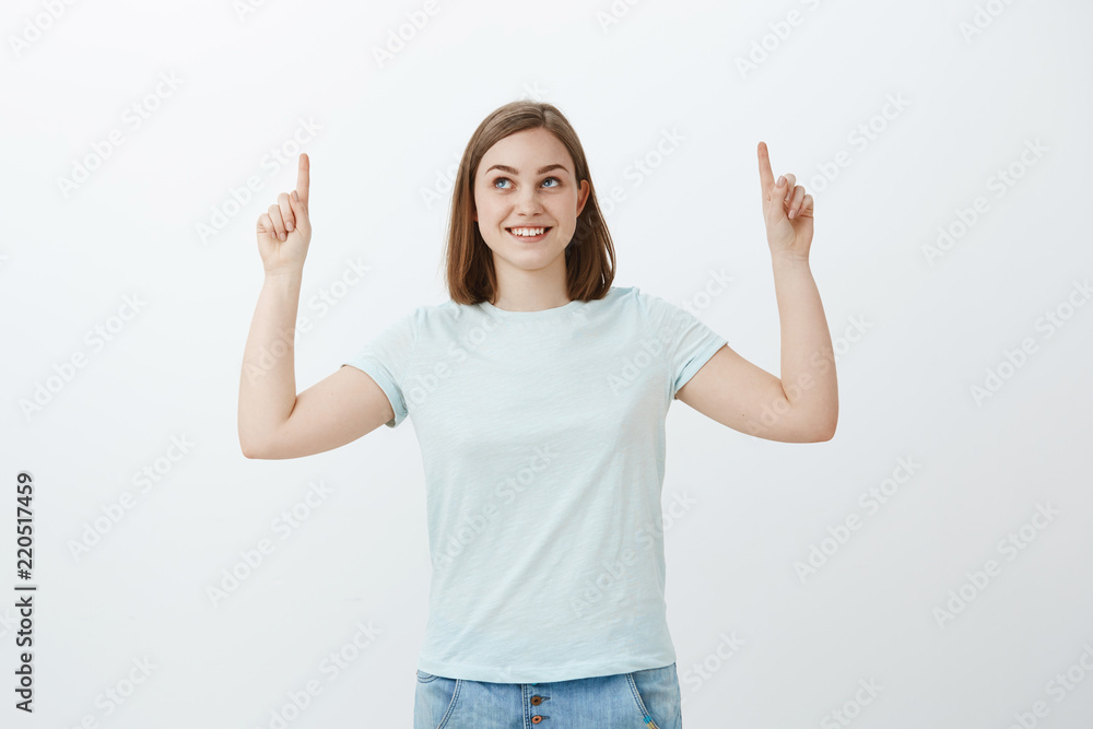 I wonder what hiding upwards. Intrigued and joyful upbeat female with broad curious smile pointing and gazing up standing in casual t-shirt and jeans over white wall promoting cool copy space I wonder what hiding upwards. Intrigued and joyful upbeat female with broad curious smile pointing and gazing up standing in casual t-shirt and jeans over white wall promoting cool copy space
