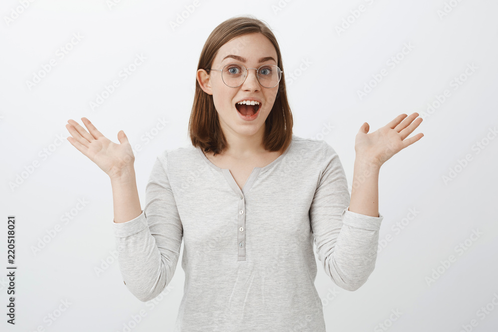 Portrait of expressive joyful smart european female in trendy glasses and white blouse talking passionately about interesting hobby gesturing with raised palms being emotional over grey wall Portrait of expressive joyful smart european female in trendy glasses and white blouse talking passionately about interesting hobby gesturing with raised palms being emotional over grey wall