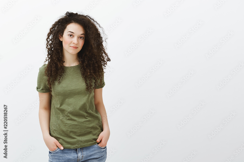 Studio shot of stylish beautiful woman with curly hair, holding hands in pockets and smiling at camera with pleased polite expression, being friendly and nice while working in flower shop Studio shot of stylish beautiful woman with curly hair, holding hands in pockets and smiling at camera with pleased polite expression, being friendly and nice while working in flower shop