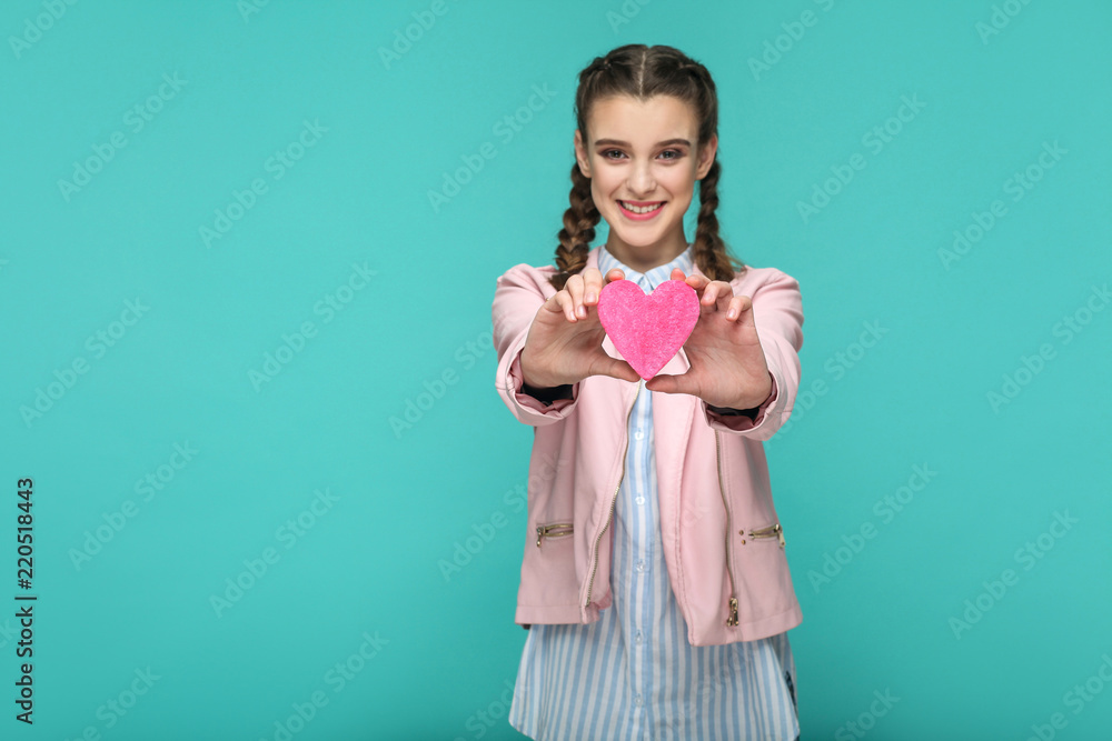 Happy beautiful girl in casual style, pigtail hairstyle and pink jacket, standing and showing pink heart shape and looking at camera with toothy smile, Indoor, isolated on blue or green background Happy beautiful girl in casual style, pigtail hairstyle and pink jacket, standing and showing pink heart shape and looking at camera with toothy smile, Indoor, isolated on blue or green background