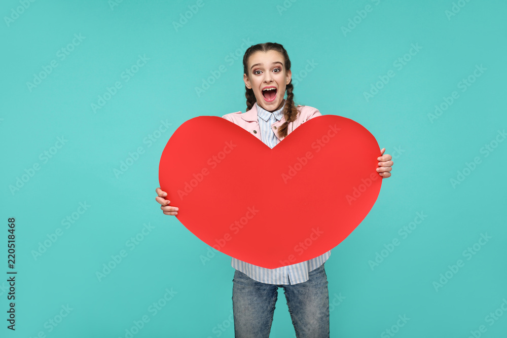 Amazed beautiful girl in casual style, pigtail hairs and jacket, standing and holding big red heart shape and looking at camera with surprised face, studio shot isolated on blue or green background Amazed beautiful girl in casual style, pigtail hairs and jacket, standing and holding big red heart shape and looking at camera with surprised face, studio shot isolated on blue or green background