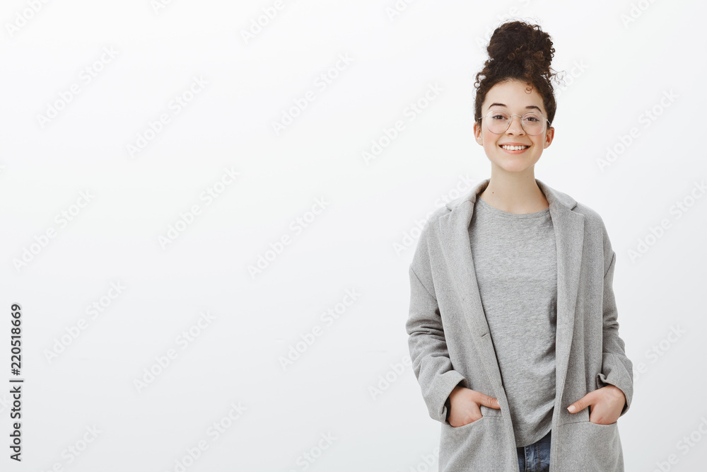 Portrait of confident happy good-looking girl with curly hair combed in bun, wearing gray coat and glasses, holding hands in pockets and smiling cheerfully at camera, feeling awesome over gray wall Portrait of confident happy good-looking girl with curly hair combed in bun, wearing gray coat and glasses, holding hands in pockets and smiling cheerfully at camera, feeling awesome over gray wall