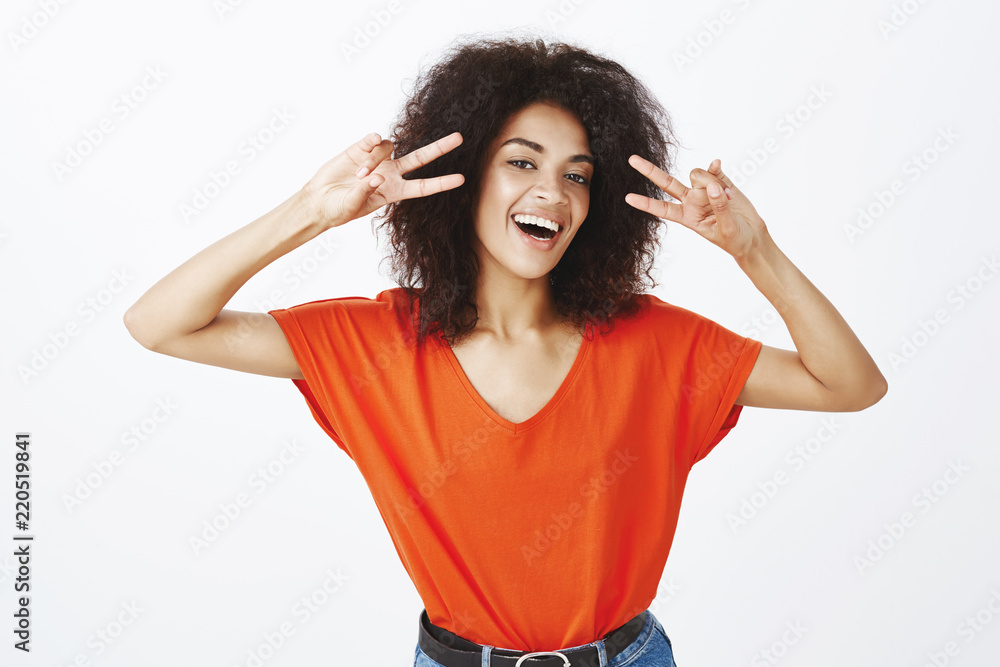 Ready to explore positive emotions of today. Happy carefree attractive woman with dark skin and afro hairstyle, smiling broadly and holding victory or peace gesture near face, standing over gray wall Ready to explore positive emotions of today. Happy carefree attractive woman with dark skin and afro hairstyle, smiling broadly and holding victory or peace gesture near face, standing over gray wall