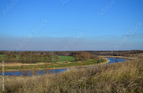 The bend of the steppe river against the backdrop of a green field. Blue sky with a haze of clouds. Dry grass in the foreground. River downhill