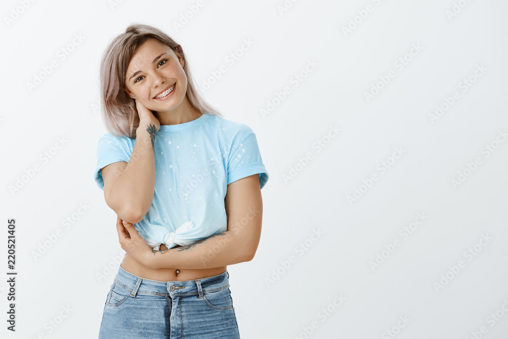 Indoor shot of feminine charming woman in stylish clothes, tilting head and smiling while touching neck and holding arm crossed over chest, being timid and unconfident standing in awkward situation Indoor shot of feminine charming woman in stylish clothes, tilting head and smiling while touching neck and holding arm crossed over chest, being timid and unconfident standing in awkward situation