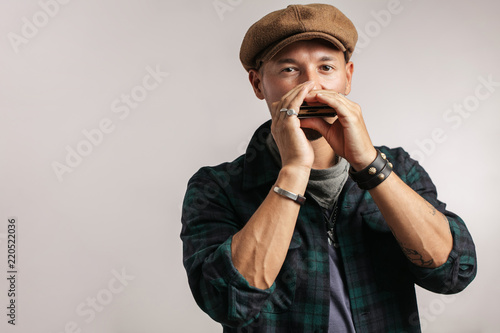 Caucasian maustached male traveler, wearing trendy cap playing a harmonica and looking at camera, isolated over white background. Travel lifestyle concept photo
