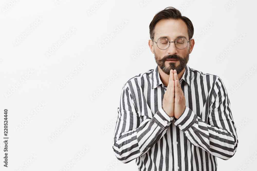 Never forgets to thank god for success. Calm and relaxed good-looking believer in glasses and striped shirt, holding hands in pray while praying or meditating with closed eyes over grey wall Never forgets to thank god for success. Calm and relaxed good-looking believer in glasses and striped shirt, holding hands in pray while praying or meditating with closed eyes over grey wall