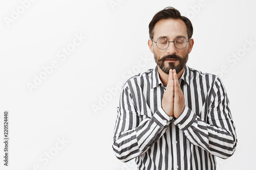 Never forgets to thank god for success. Calm and relaxed good-looking believer in glasses and striped shirt, holding hands in pray while praying or meditating with closed eyes over grey wall