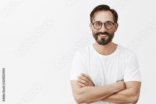At your service. Charming carefree and confident european man with beard and dark hair in glasses and white t-shirt, holding hands crossed on chest in self-assured pose, smiling joyfully at camera photo