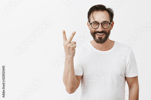 Indoor shot of beautiful confident and happy man with trendy glasses showing number two or up yours gesture, smiling broadly over gray background, making something twice times photo