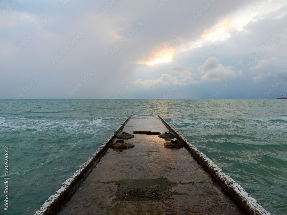 Concrete breakwater with light reflection on a wet surface, turquoise sea under the sky with dramatic clouds in a small storm Concrete breakwater with light reflection on a wet surface, turquoise sea under the sky with dramatic clouds in a small storm