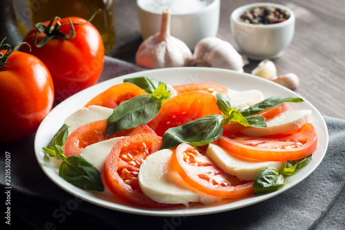 Photo of Caprese Salad with tomatoes, basil, mozzarella, olives and olive oil on wooden background. Italian traditional caprese salad ingredients. Mediterranean, organic and natural food concept.