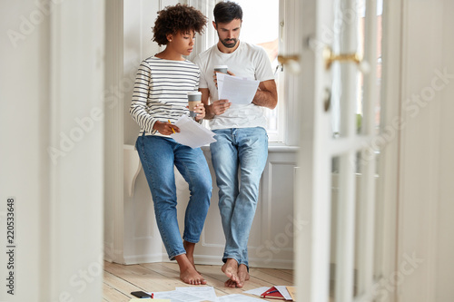 Full length shot of African American woman and her business partner work on common business project, study graphics in papers, meet in informal situation, pose at apartment, dressed casually © wayhome.studio 