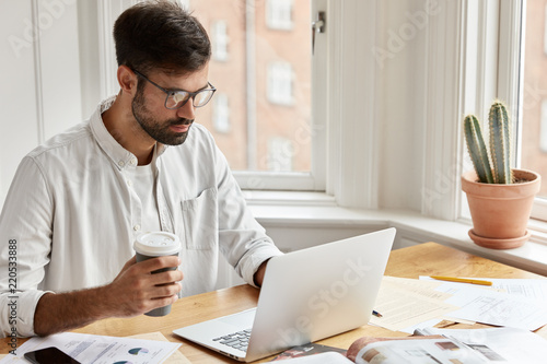 Concentrated male executive manager focused into screen of laptop computer, drinks takeway coffee, works on accounting report, does paperwork, wears glasses and white shirt, sits at workplace