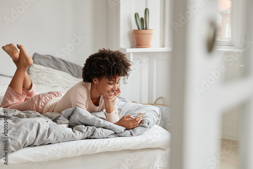Cheerful teengae girl lies in bed, uses mobile phone, recieves text message from boyfriend, wears nighclothes, poses in spacious cozy bedroom, feels relaxed. People, relaxation, technology concept