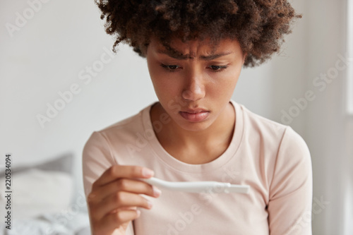 Desperate sad unhappy black woman looks at pregnancy test, being discontent with result, isnt ready to have baby, poses against domestic interior. People, ethnicity, maternity and fertility concept