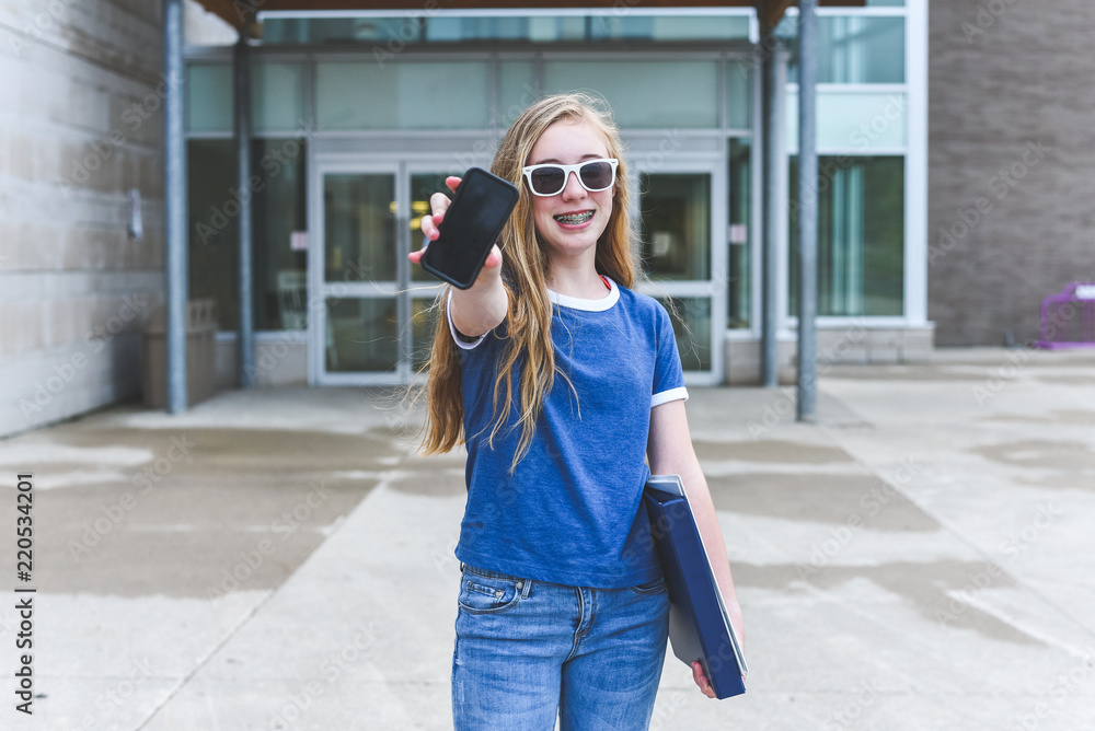 Happy teenage girl showing off cell phone while standing in front of a school.