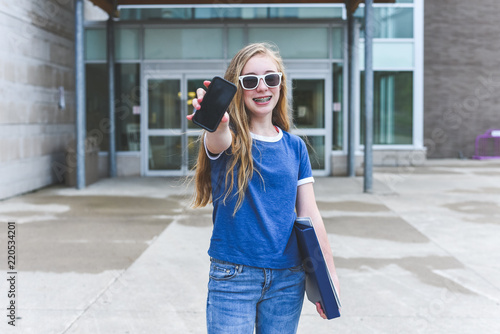 Happy teenage girl showing off cell phone while standing in front of a school. © Brian