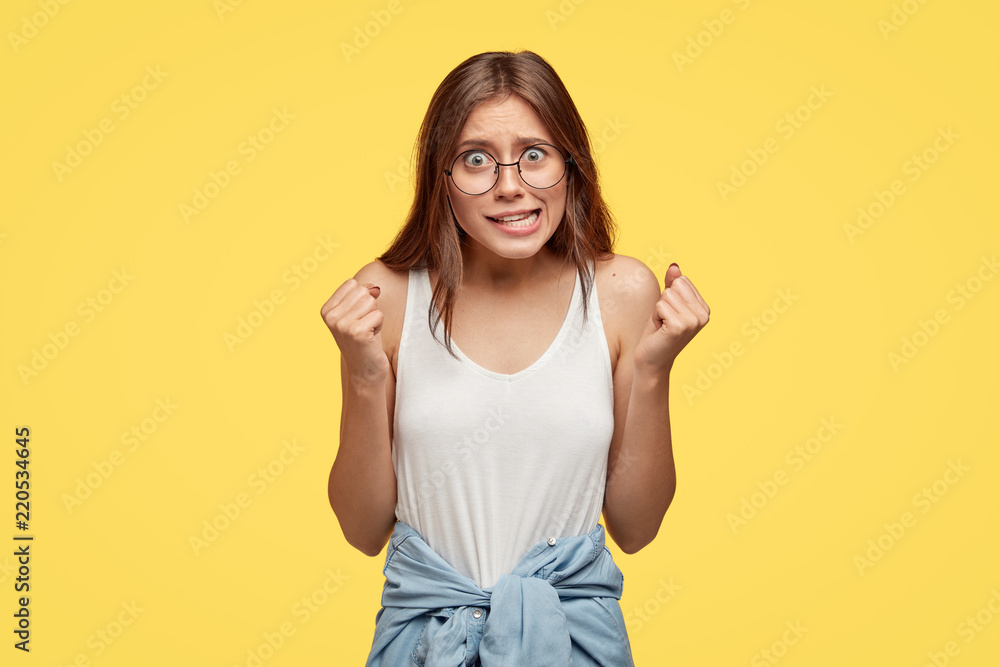 Powerful model tries to control emotions, clenches fists while anticipates announcement of results, looks wth great desire at camera, stands against yellow studio wall, expresses her feelings Powerful model tries to control emotions, clenches fists while anticipates announcement of results, looks wth great desire at camera, stands against yellow studio wall, expresses her feelings