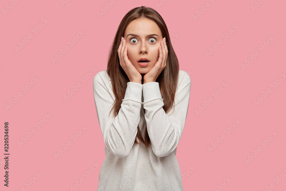 Bugged eyed emotional young woman keeps hands on cheeks, stares in terror, dressed in casual clothes, poses against pink wall, hears bad news about close person. Stupefied teenager stands indoor Bugged eyed emotional young woman keeps hands on cheeks, stares in terror, dressed in casual clothes, poses against pink wall, hears bad news about close person. Stupefied teenager stands indoor