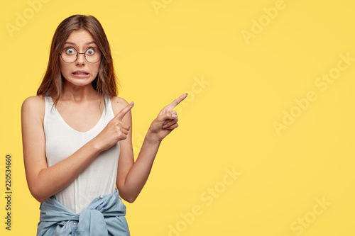 Oops, come and look there! Lovely young woman feels worried and ashamed, points with both index fingers at upper right corner, isolated over yellow background, makes mistake and looks awkward