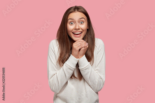 People, unexpectedness and emotions concept. Overjoyed European woman clasps hands from happiness, has broad smile and eyes full of delight, dressed in white sweatshirt, stands indoor alone.