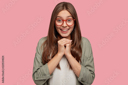 Studio shot of pleased lovely human keeps hands together under chin, being in high spirit, likes friends idea for next day, wears spectacles with red rim, isolated on pink wall. Positive emotions